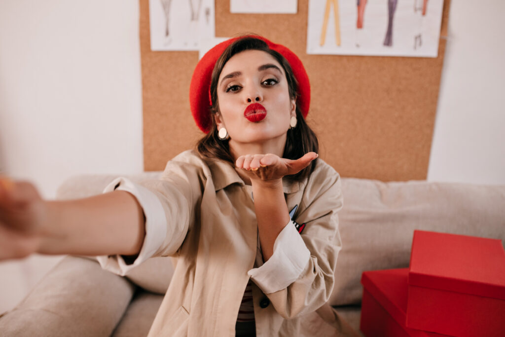 Stylish lady in beige stylish trench blows kiss and takes selfie. Modern young woman takes photo an